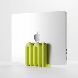 Puede incluir: Un ordenador portátil plateado con acabado metálico y logotipo de Apple se mantiene en posición vertical mediante un soporte de silicona en forma de U de color verde lima. El soporte tiene tres secciones tubulares redondeadas que sujetan.