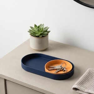 Op de afbeelding: Een marineblauw ovaal dienblad met een kleinere ronde oranje schaal waarin een metalen sleutelbos ligt. Achter het dienblad staat een kleine groene vetplant in een lichtbeige keramische pot op een vlakke ondergrond.