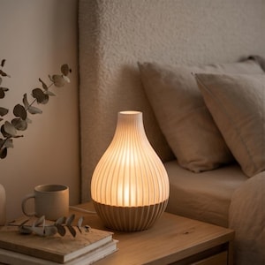 Może przedstawiać: Biała, gruszkowata lampa stołowa o żebrowanej, pionowo prążkowanej powierzchni, emitująca ciepłe, delikatne światło. Lampa zapewnia nastrojowe oświetlenie szafki nocnej w otoczeniu o neutralnej kolorystyce i miękkich teksturach.