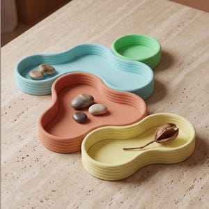 Op de afbeelding: Een set van drie onregelmatige, golvende schalen in lichtblauw, terracotta en lichtgeel, vergezeld door een kleine ronde groene schaal. De schalen hebben geribbelde zijkanten en bevatten gladde rivierstenen en een gedroogde zaadpeul als decoratie.