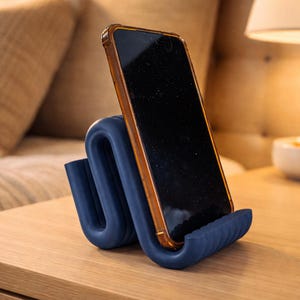 Può includere: Un supporto per telefono in silicone blu scuro a forma di S con una consistenza a coste, che sostiene uno smartphone con schermo scuro e cornice marrone. Il supporto presenta un design curvo che mantiene il dispositivo in posizione verticale.