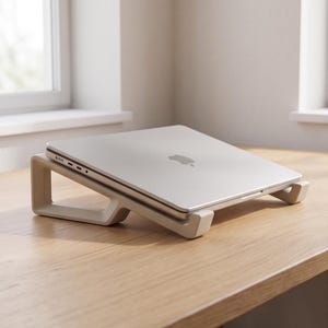 Peut inclure: Un ordinateur portable argenté posé sur un support incliné de couleur beige clair. Le support présente une structure géométrique évidée qui surélève l'appareil. L'ordinateur est fermé, affichant une finition métallique et un logo central.