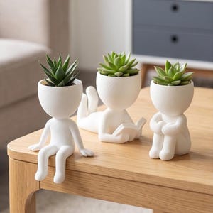 Op de afbeelding: Drie witte, matte keramische plantenbakken in de vorm van menselijke figuren, elk met een groene vetplant. Eén figuur zit met bungelende benen, één ligt op de buik een boek te lezen en één zit met opgetrokken knieën op een houten oppervlak.