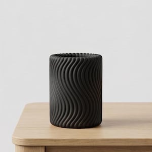 Può includere: Un vaso cilindrico nero con una superficie ondulata e strutturata. Il vaso è realizzato in un materiale opaco e poggia su una superficie di legno chiaro. Il bordo superiore presenta un design a coste.