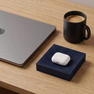 Peut inclure: Un plateau carré bleu foncé avec une texture de surface ondulée et nervurée contient un boîtier de charge blanc pour écouteurs sans fil. Le plateau est placé à côté d'un ordinateur portable gris fermé et d'une tasse en céramique noire mate.
