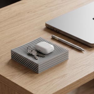 Op de afbeelding: Een grijze, rechthoekige tray met een geribbeld oppervlak bevat een set sleutels en een witte draadloze oortelefoonhoes. Een zilveren stylus en een laptop staan op de achtergrond. De tray staat op een houten bureau.