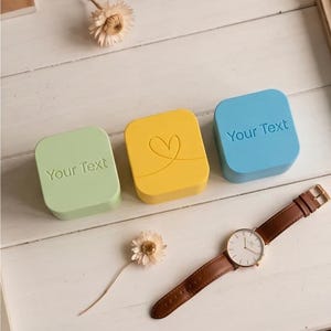 Pode incluir: Três caixas quadradas em verde claro, amarelo e azul claro. A caixa verde tem o texto Your Text, a caixa amarela apresenta um contorno de coração e a caixa azul tem o texto Your Text. Um relógio de pulso com pulseira de couro castanho está incluído.