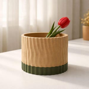 May include: A cylindrical vase with a textured, light brown upper section and a dark green lower band. A single red tulip with green leaves is placed inside the vase. The vase is on a white surface.