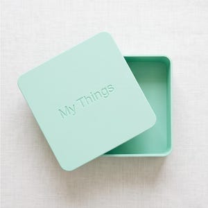 May include: A square mint green storage box with a matching lid. The lid features the embossed text "My Things" in a simple sans-serif font. The box is open, revealing a hollow interior, and the lid is placed at an angle on top of the base.