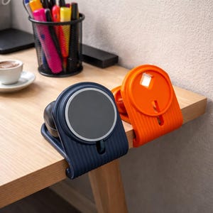 Op de afbeelding: Twee bureau-organizers worden getoond. Eén is marineblauw met een grijze cirkelvormige bovenkant en de andere is feloranje. Beide zijn vastgeklemd aan de rand van een houten bureau. Een zwarte pennenhouder van gaas en een kopje koffie staan op de achtergrond.