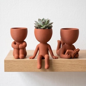 Op de afbeelding: Drie terracotta-kleurige plantenbakken in de vorm van menselijke figuren met een textuur. De linkerfiguur zit met opgetrokken knieën, de middelste figuur zit rechtop met bungelende benen en de rechterfiguur ligt neer en leest een boek.