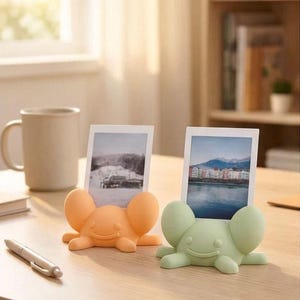 Könnte beinhalten: Zwei kleine, abgerundete Fotohalter in Form von Kreaturen in Pastellorange und Hellgrün. Jeder Halter hat ein lächelndes Gesicht und breite, ohrenartige Vorsprünge, um ein vertikales, rechteckiges Foto zu halten. Der orangefarbene Halter zeigt eine Schneelandschaft, der grüne eine Stadtansicht am Wasser.