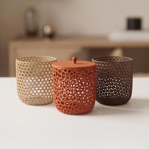 Può includere: Tre contenitori cilindrici con complessi motivi a reticolo traforato. Il contenitore a sinistra è beige, quello centrale è arancione terracotta con coperchio abbinato e quello a destra è marrone scuro. Ogni vaso presenta una struttura cava e porosa.