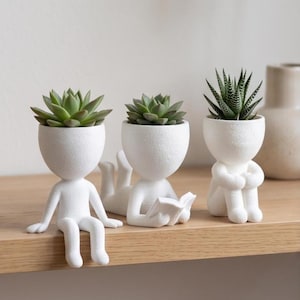 May include: Three white matte ceramic planters shaped like human figures, each holding a green succulent. One figure sits with legs dangling, one lies on its stomach reading a book, and one sits with knees pulled to its chest in a seated position.