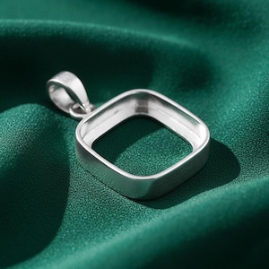 May include: A silver pendant setting with a square bezel and a loop for a chain. The pendant is made of a shiny, metallic material and is displayed against a dark green fabric background.
