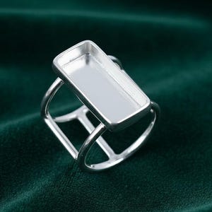 May include: A silver-toned ring with a rectangular bezel setting. The ring features a modern design with a rectangular top and a unique, openwork band. The setting is designed to hold a rectangular stone or embellishment.