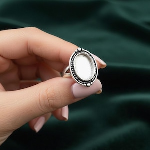 Könnte beinhalten: Ein silberner Ring mit einer ovalen Lünettenfassung. Der Ring hat eine dekorative, seilartige Umrandung um die Lünette und kleine Perlenakzente. Der Ring wird von einer Hand mit lackierten Nägeln gehalten.