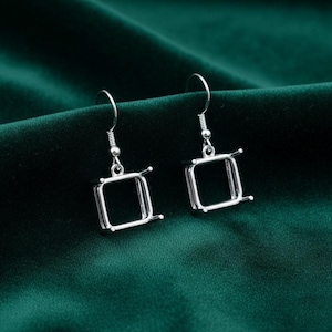 May include: A pair of silver square drop earrings. Each earring has a square frame with four small prongs at each corner, suspended from a curved ear wire. The earrings are set against a dark green backdrop.