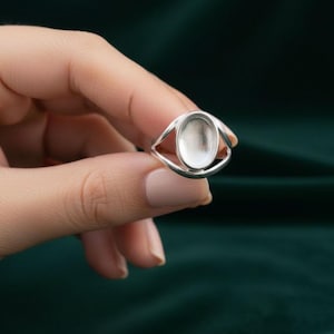May include: A silver ring with an oval setting, held by a hand. The ring's design features a sleek, minimalist style with a polished finish. The setting is designed to hold a stone or other decorative element.