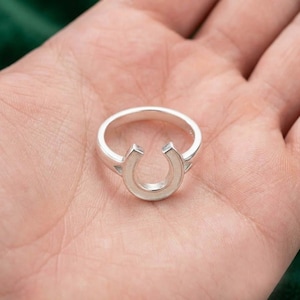 Könnte beinhalten: Ein silberfarbener Ring mit einem hufeisenförmigen Mittelstück. Die Ringschiene ist glatt und kreisförmig und geht in das nach oben geöffnete Hufeisen-Design über. Das Metall weist durchgehend eine polierte und reflektierende Oberfläche auf.