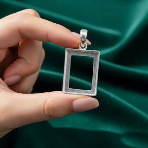 May include: A silver-colored rectangular pendant with a hollow center, held against a dark green fabric background. The pendant has a loop at the top for a chain. The pendant is held between two fingers.