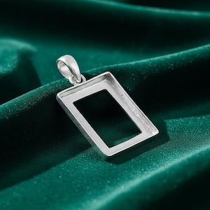 May include: A silver rectangular pendant with a loop at the top. The pendant is set against a dark green, velvety background. The pendant's frame is empty, designed to hold a stone or other decorative element.