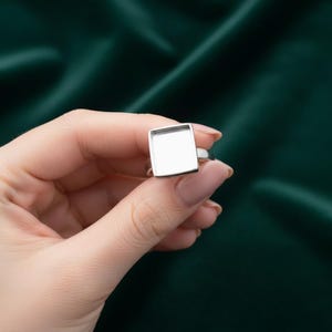 Op de afbeelding: Een zilveren ring met een vierkante bezel, gehouden tegen een donkergroene achtergrond. De zetting van de ring is een leeg vierkant, klaar voor personalisatie. De ring wordt in een close-up getoond, waarbij de gepolijste metalen afwerking wordt benadrukt.