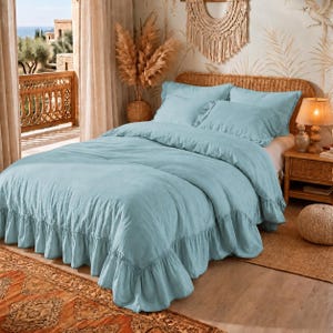 May include: A light blue bedding set featuring a duvet cover and two matching pillowcases with ruffled edges. The fabric has a visible textured weave, and the duvet cover drapes over the sides of the bed with a wide, gathered ruffle trim.