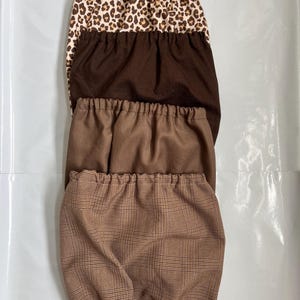 Peut inclure: Une pile de quatre housses en tissu avec des bords élastiqués. La housse supérieure présente un imprimé léopard beige et noir, suivie d'une housse marron foncé unie, d'une housse marron moyen unie et d'une housse à motif écossais marron et noir.