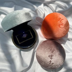 May include: A silver ring with a central stone sits in an open light blue velvet box. Beside it are two closed round velvet boxes, one orange with the embossed letters M&E and one mauve with the embossed letters M&M.