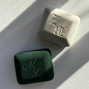 Pode incluir: Duas caixas de joias retangulares de veludo, uma em cinza claro com as letras ZE em relevo numa fonte gótica, e outra em verde floresta escuro com as letras cursivas ML em relevo. Ambas as caixas apresentam um acabamento suave e cantos arredondados.