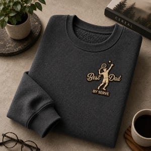 Könnte beinhalten: Dunkelgraues Sweatshirt mit dem Text "Best Dad" und einer Tennisspieler-Grafik. Das Sweatshirt hat einen Rundhalsausschnitt und gerippte Bündchen. Der Text "BY SERVE" befindet sich unter der Grafik.