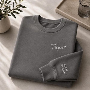 Könnte beinhalten: Ein anthrazitfarbenes Sweatshirt mit Rundhalsausschnitt und weißer Stickerei auf der Brust, die das Wort Papa und ein kleines Herz zeigt. Am linken Ärmelbündchen sind die Namen LIAM und NOAH in Weiß sowie ein kleines Herz eingestickt.