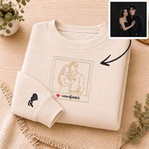 May include: A beige crewneck sweatshirt featuring a central embroidered line art portrait of a couple inside a square frame, accompanied by a small red heart and a sound wave graphic. A black heart icon is embroidered on the left sleeve cuff.