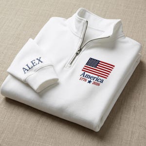 May include: A white quarter-zip pullover featuring an embroidered American flag with the text America 1776 2026 below it. The sleeve cuff is embroidered with the name ALEX in navy blue thread. The garment has a silver-toned metal zipper pull.