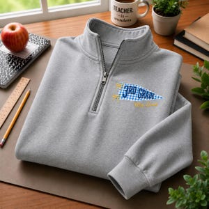 Könnte beinhalten: Ein hellgrauer Pullover mit kurzem Reißverschluss und einer gestickten blau-weiß karierten Wimpel-Applikation auf der Brust. Der Wimpel zeigt den Schriftzug 3RD GRADE in blauen Buchstaben und den Namen Mrs. Louis in gelber Schreibschrift.