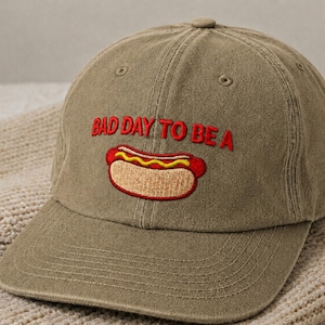 May include: Olive green baseball cap with the embroidered phrase "BAD DAY TO BE A" above a hot dog graphic. The hot dog is red with yellow mustard and a textured bun. The cap has a curved brim and a button on top.