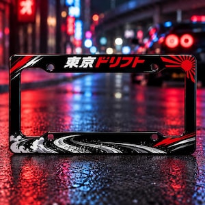 Può includere: Un portatarga nero con il testo giapponese Tokyo Drift in bianco e rosso. Il design include un sole nascente rosso sul lato destro, fumo di pneumatici bianco stilizzato e una grafica stradale nera lungo il bordo inferiore.