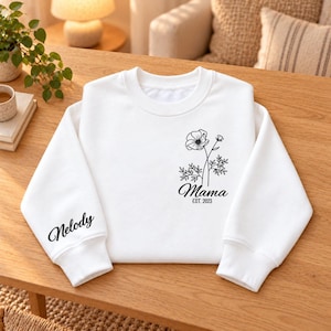 May include: A white long-sleeve sweatshirt featuring a black line art floral design on the left chest with the text Mama EST. 2023 underneath. The left sleeve displays the name Nelody in a black cursive script font on the lower portion.
