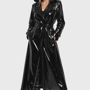 Könnte beinhalten: Ein langer, schwarzer, glänzender Trenchcoat aus lackartigem Material. Der Mantel verfügt über eine Kapuze, eine zweireihige Knopfleiste auf der Vorderseite und einen passenden Bindegürtel in der Taille. Er hat lange Ärmel mit Schnallen.