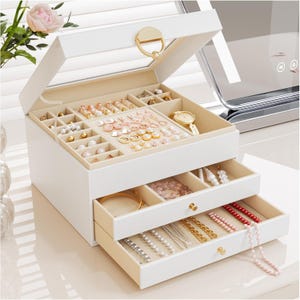 Op de afbeelding: Een witte sieradendoos met een glazen deksel, een gouden ronde sluiting en twee uitschuifbare lades. De binnenkant is bekleed met beige fluweel en bevat diverse vakjes voor oorbellen, ringen, kettingen, armbanden en een gouden polshorloge.