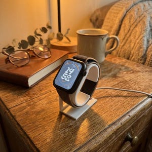 Peut inclure: Une montre connectée noire avec un bracelet gris foncé est posée sur un support de chargement blanc. L'écran affiche 14h52. Le support est sur une table de chevet en bois, à côté d'un livre, de lunettes et d'une tasse à café.