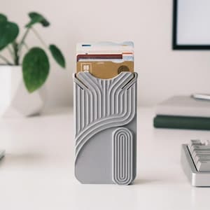 May include: A grey card holder with a distinctive, curved pattern, holding several cards. It stands on a white desk. A small plant in a white geometric pot and a white cup are visible in the background.