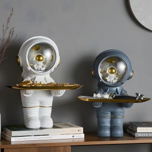 Könnte beinhalten: Zwei dekorative Astronauten-Skulpturen, eine weiße und eine grau-blaue. Jeder Astronaut hält ein goldfarbenes Tablett. Im Inneren des Helms befindet sich eine kleine Astronautenfigur und goldfarbene Akzente.