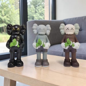 May include: Three KAWS-style figurines in black, grey-green, and brown. Each figure has crossed-out eyes and holds a bundle of green paper. The figures stand on a light-coloured wooden table.