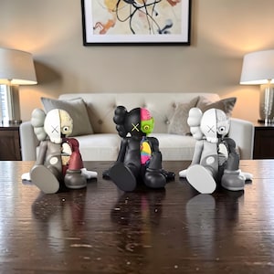 May include: Three seated KAWS Companion figures with exposed internal anatomy. The figures are in shades of gray, black, and a vibrant black with a green mask. The figures have the signature X eyes and are displayed on a dark wooden surface.