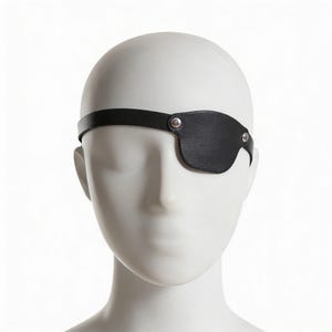 Könnte beinhalten: Eine schwarze Augenklappe aus Leder mit einem Riemen, befestigt mit silberfarbenen Nieten, wird auf einem Schaufensterkopf gezeigt. Die Augenklappe ist so konzipiert, dass sie ein Auge bedeckt und aus einem strukturierten Material besteht.