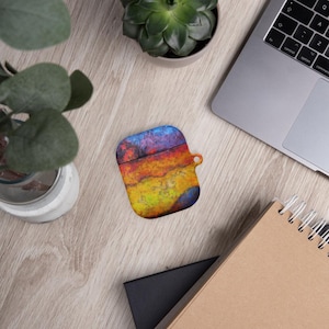 Könnte beinhalten: Eine farbenfrohe AirPod-Hülle mit einem lebendigen, abstrakten Design. Die Hülle weist eine Mischung aus Blau-, Rot-, Orange- und Gelbtönen auf, die ein strukturiertes, künstlerisches Aussehen erzeugt. Die Hülle hat eine kleine Schlaufe zum Anbringen an einem Schlüsselanhänger oder einer Lanyard. Die Hülle liegt auf einer Holzoberfläche.