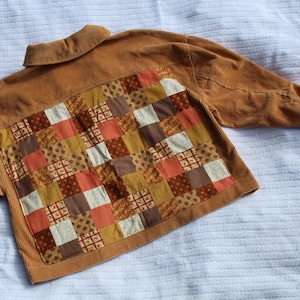 May include: A tan corduroy jacket featuring a large rectangular back panel composed of a patchwork design. The patchwork consists of various square fabric pieces in shades of brown, orange, cream, and mustard yellow with floral patterns.