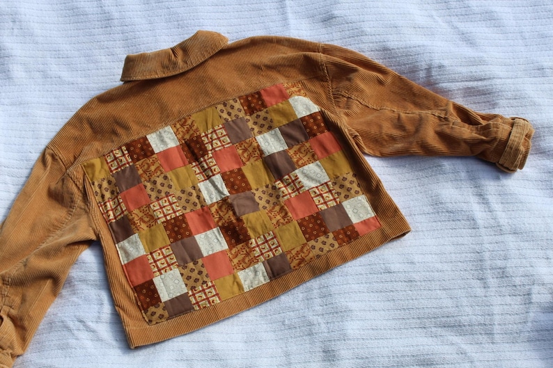 May include: A tan corduroy jacket featuring a large rectangular back panel composed of a patchwork design. The patchwork consists of various square fabric pieces in shades of brown, orange, cream, and mustard yellow with floral patterns.